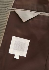 Brauner Stoff mit einer glatten Textur, versehen mit einem beigen, dreieckigen Klappen und einem quadratischen weißen Patch mit einem Gittermuster.