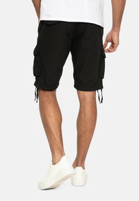 Homme portant un short cargo noir et des baskets blanches, vu de dos sur un fond uni.