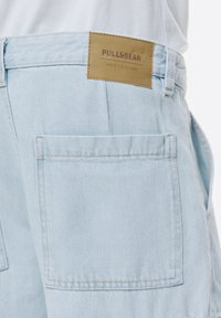 Vaaleansiniset farkkushortsit, joissa on takana ruskea etiketti, jossa lukee "PULL&BEAR DENIM COLLECTION". Sisältää takataskun, jossa on näkyvä ommel.