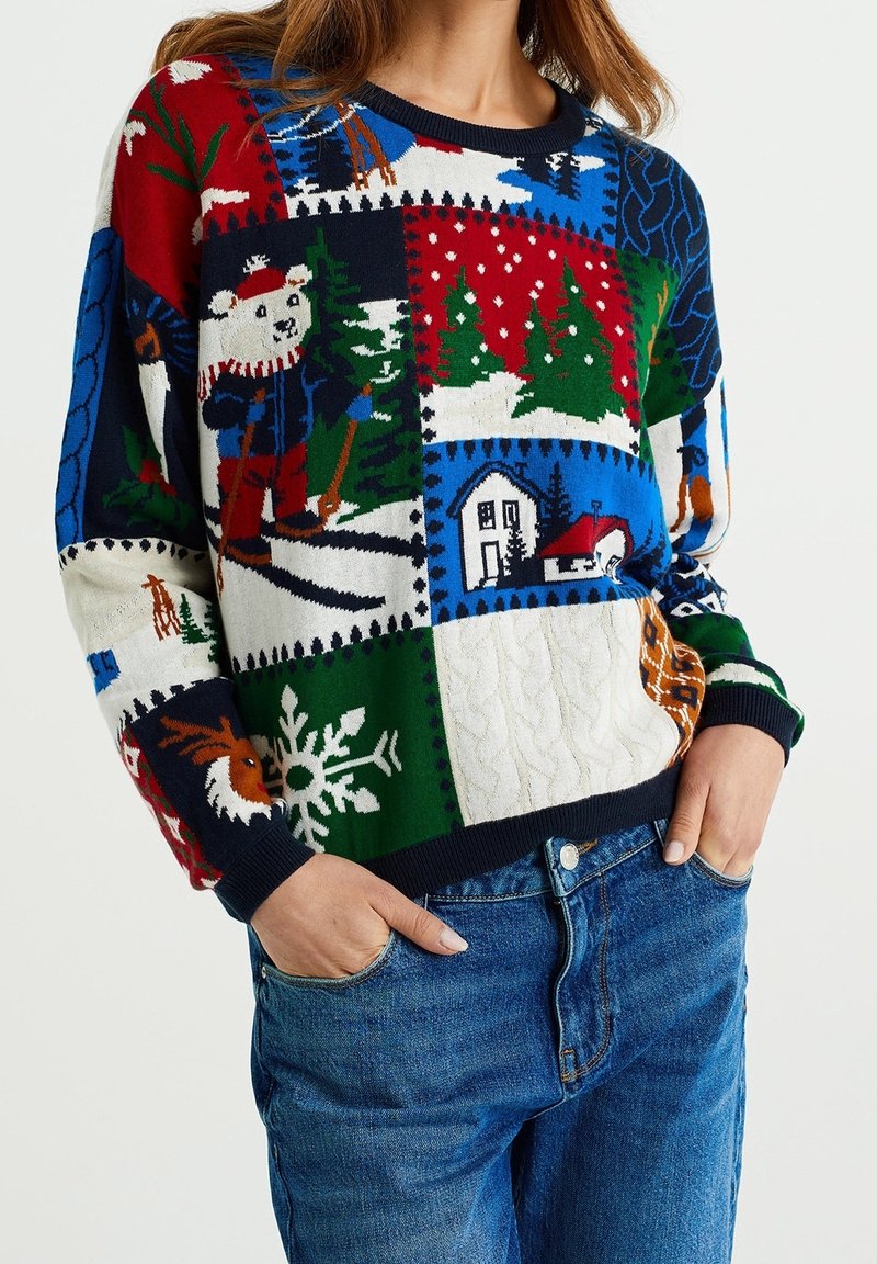 Femme portant un pull de Noël patchwork coloré avec un ours polaire, des pins, des flocons de neige, des maisons et des rennes, associé à un jean bleu.