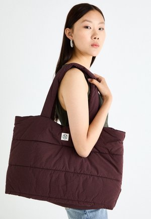 Bolso tote de color burdeos oscuro con textura acolchada, que cuenta con asas anchas y un pequeño parche con el logotipo. La tela parece suave y acolchada.
