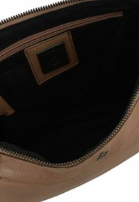 Interno di una borsa in pelle marrone con fodera nera, dotata di chiusura con zip e una patch in pelle color nocciola con logo. Texture liscia e forma arrotondata.