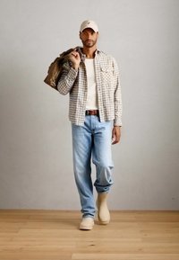 Chemise à carreaux beiges sur un pull blanc, jeans bleu clair et chaussures beiges ; tenant un sac marron ; fond neutre et sol en bois.