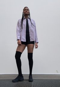 Chemise large rayée violet clair, bodysuit noir et chaussettes noires hosières, accompagnés de chaussures de sport noires, se tenant sur une surface texturée.
