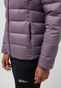 Giacca imbottita viola con sezioni trapuntate, tasca con zip e una texture liscia, indossata sopra pantaloni neri con logo.
