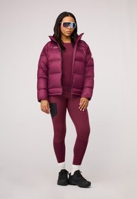 Burgundowa kurtka puchowa o połysku, w zestawie z dopasowanym topem i legginsami z czarnymi akcentami oraz czarnymi butami sportowymi.