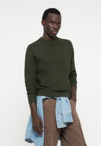 Pulover verde tricotat, cu guler rotund, asortat cu pantaloni cargo cărămizii. O cămașă din denimul albastru deschis este legată în jurul taliei.