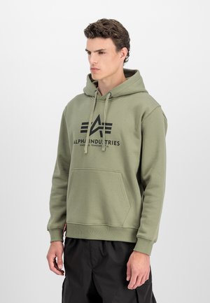Jeune homme portant un hoodie vert d'Alpha Industries avec une poche à l'avant et un pantalon noir, se tenant devant un fond clair.
