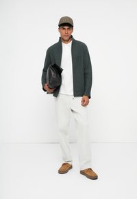 Jacket de polar verde com fecho éclair, t-shirt branca, calças claras e sapatos castanhos. Segura uma mala preta e usa um boné xadrez.