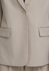 Blazer beige sur mesure avec un seul bouton, deux poches latérales et des revers crantés, fabriqué dans un tissu lisse et structuré.