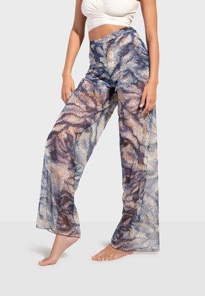 Vrouw draagt hooggetailleerde, wijde transparante broek met blauw, beige en bruin abstract patroon, gecombineerd met een wit cropped topje.