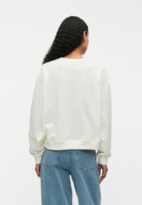 White sweatshirt with a crew neck and relaxed fit, featuring ribbed cuffs and hem. Worn with light blue, loose-fit jeans.