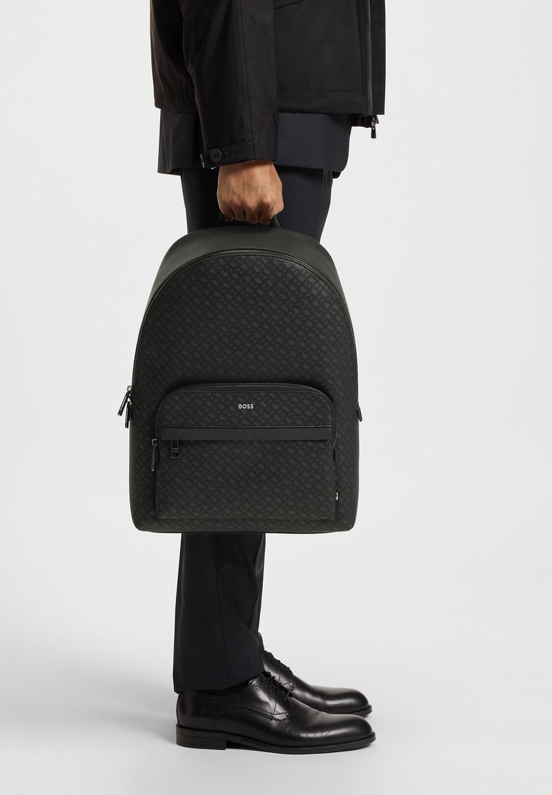 Sac à dos noir texturé avec une forme arrondie, doté d'une poche zippée à l'avant et d'un détail de logo, tenu par une personne en tenue professionnelle.