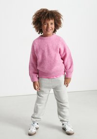 Ružový pletený sveter s textúrovaným povrchom, skombinovaný so svetlošedými teplákmi a bielymi teniskami. Dieťa stojí sebavedomo a usmieva sa.