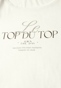 Hvid bomulds t-shirt med trykt tekst i varierede skrifttyper: "Le TOP DU TOP OWN THE DAY" og et slogan nedenfor i en lysere nuance.