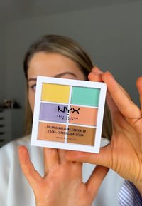 La palette de correcteurs colorés NYX dans un boîtier blanc présente six carrés : des teintes jaune, verte, lavande, pêche et marron.