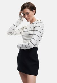 Gestreepte witte trui met zwarte lijnen, lange mouwen en een strakke zwarte rok van textiel. Model poseert met hand op hoofd.