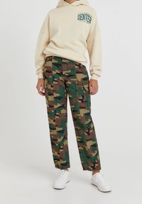 Beige hoodie met "DENVER" opdruk, gecombineerd met groen-bruin camouflage cargobroek met zakken, en witte sneakers.