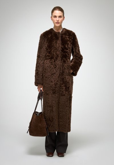 Bruine jas van nepbont met een hoge kraag, lange mouwen en een open voorkant; gecombineerd met een bruine suède handtas en donkere, op maat gemaakte broek.