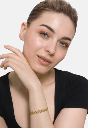 Gouden armband met ingewikkeld bloemenontwerp, gedragen om de pols. Model heeft een lichte huid, draagt een zwarte top en toont een elegante handhouding.