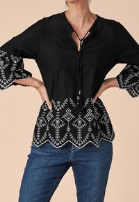 Blouse noire avec des motifs brodés blancs à l'ourlet et aux manches. Dotée d'un col en V avec des liens et d'une coupe décontractée. Associée à un jean bleu.