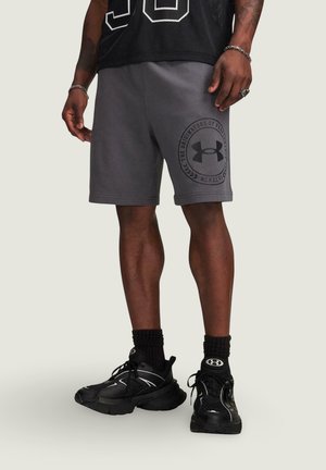 Mężczyzna noszący ciemnoszare szorty Under Armour z logo na lewej nodze, czarne skarpety i czarne buty sportowe, stojący na jednolitym tle.