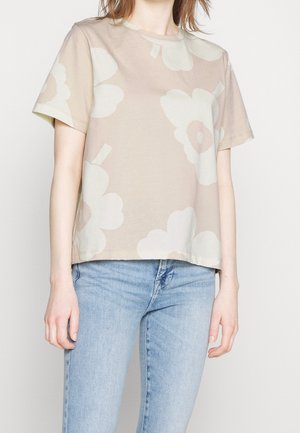 Femme portant un T-shirt beige à manches courtes avec un grand motif floral blanc et un jean bleu clair, debout devant un fond clair uni.
