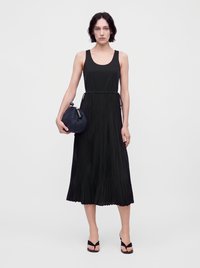 Woman wears sleeveless black pleated midi dress and black open-toe sandals, holding a small dark blue textured clutch bag.