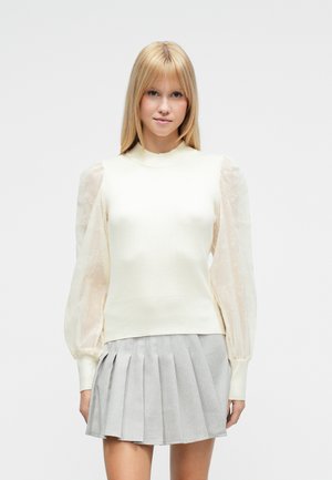 Femme blonde portant un pull crème avec des manches bouffantes transparentes et une mini-jupe plissée gris clair, debout devant un fond blanc.
