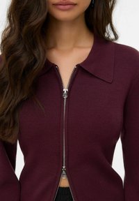 Bordeaux kleurige zip-up trui met een brede kraag, geribbelde textuur en een zilveren rits. Draagt zich met zwarte stof eronder, wat het getailleerde ontwerp accentueert.