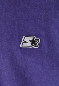 Violet medvilnės audinys su balto krašto siuvinėtu logotipu, kuriame matomas juodas žvaigždutė ir stilizuota "S" kairiajame apatiniame kampe.