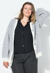 Cardigan grigio e bianco a fantasia con chiusura a bottoni, polsini a coste e vestibilità rilassata, indossato sopra una maglietta nera e pantaloni a righe.
