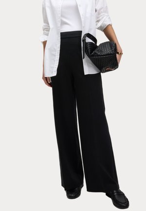 Femme portant une chemise blanche, un top blanc, un pantalon noir à jambes larges, des mocassins noirs, tenant un sac à main noir tissé avec une sangle à boucle.