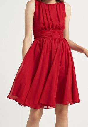 Robe rouge sans manches, longueur genou, avec une ceinture froncée et une jupe fluide plissée portée par une personne debout.