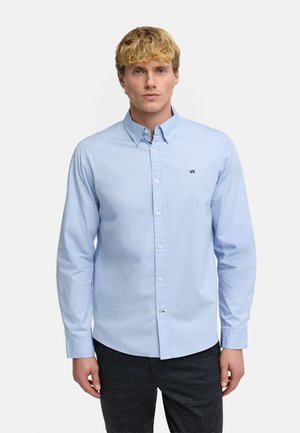 Lyshåret mand iført en lyseblå skjorte med knapper og en lille sort logo på brystet samt mørke ternede bukser, der står mod en ensfarvet baggrund.