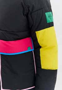 Čierna prešívaná bunda so žiarivo žltou časťou rukávov, panelmi v ružovej a červenej farbe, modrým lemom a zelenou nášivkou s nápisom "Farebná revolúcia."