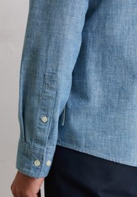 Chemise en denim bleu clair avec un poignet boutonné, présentant une texture de tissage fin et des boutons blancs contrastants. Comprend une étiquette sur le côté.