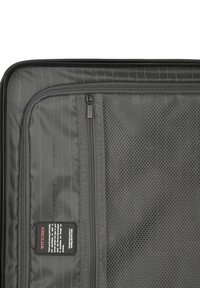 L'intérieur de la valise en tissu noir présente des poches en maille, des compartiments zippés et un motif rayé, avec une étiquette de marque affichée de manière proéminente.