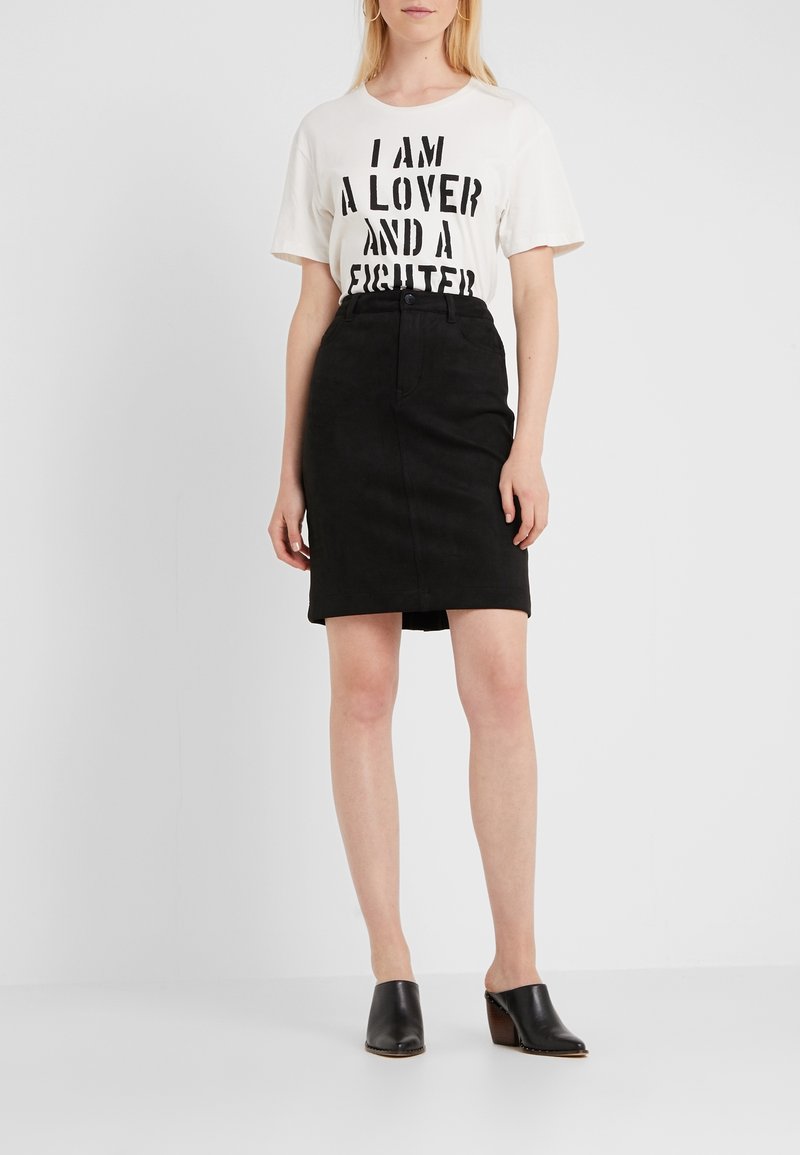 Vit t-shirt med svart text som säger "JAG ÄR EN ÄLSKARE OCH EN KÄMPARE," ihop med en svart denimkjol av medellängd och svarta klackskor.