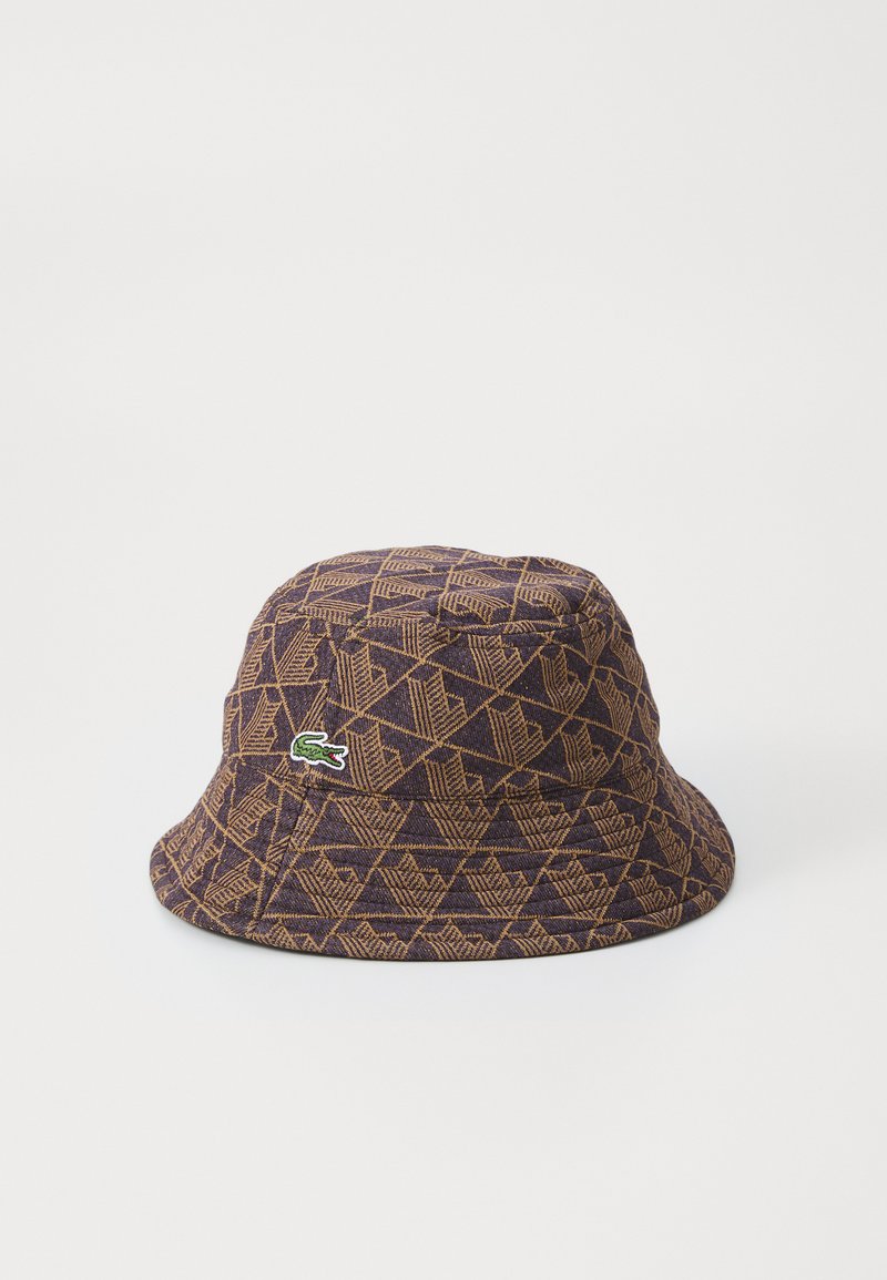 Gorra tipo bucket de tela púrpura oscura con un patrón geométrico en marrón claro. Presenta un pequeño logo verde de Lacoste en el lateral.