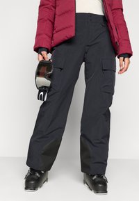 Calças de neve pretas com bolsos cargo, tecido à prova de água e pernas largas, combinadas com botas de esqui pretas. Um par de óculos de esqui é segurado em uma mão.