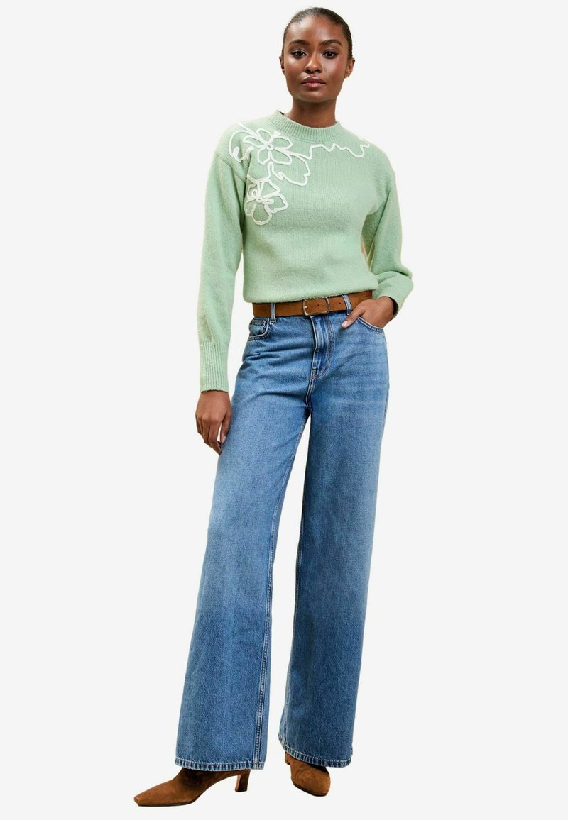 Pulover tricotat verde mentă cu broderie florală albă, asortat cu blugi largi din denim albastru și cizme cu toc mediu de culoare maro.