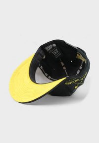 Musta lippalakki keltaisella lieriöllä, joka on varustettu tekstuuripinnalla ja brodeeratuilla yksityiskohdilla etupaneelissa. Sisäpuolella näkyy painetut tarrat.