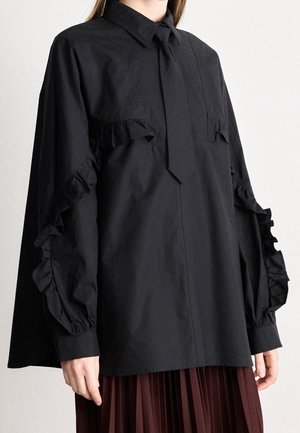 Frau trägt eine lockere schwarze Bluse mit Rüschenakzenten an Ärmeln und Brust, kombiniert mit einem plissierten dunkelbraunen Rock.