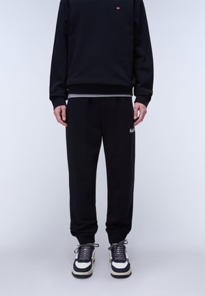 Man draagt een zwarte trui en bijpassende joggingbroek met logo op de dij, gecombineerd met witte en zwarte sneakers tegen een effen achtergrond.