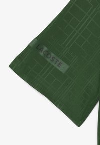 Tessuto verde scuro con un sottile motivo geometrico e un'etichetta in gomma che recita "LACOSTE" vicino al bordo.