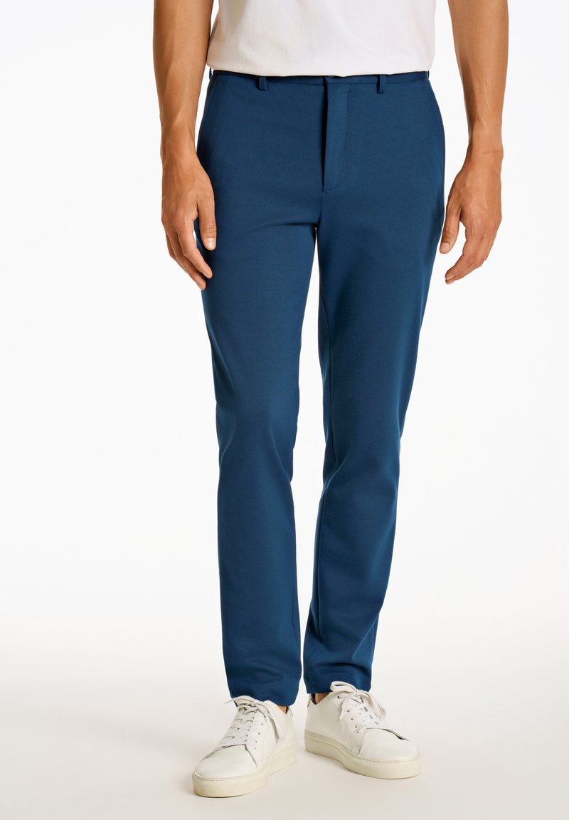 Blaue Hosen mit tailliertem Schnitt, aus einem glatten, dehnbaren Stoff gefertigt. Getragen mit weißen Turnschuhen, die eine flache Vorderseite und seitliche Taschen haben.