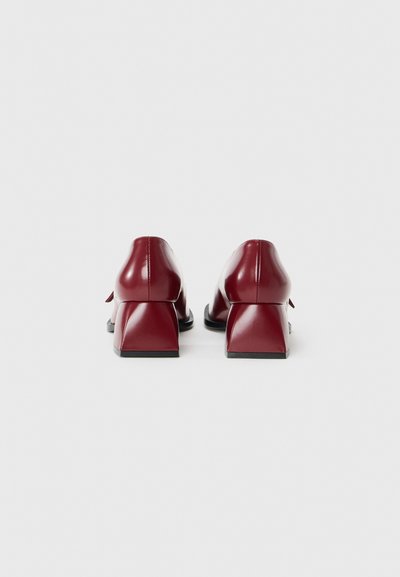 Talons en cuir bordeaux avec une forme de bloc géométrique et des semelles noires. Texture lisse avec un minimum de détails, ornés d'un accent de nœud à l'arrière.