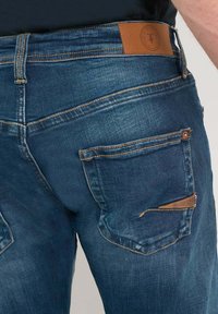 Jeans de mezclilla en azul oscuro con un acabado texturizado. Las características incluyen un parche de cuero marrón claro en la cinturilla y costuras decorativas en los bolsillos.