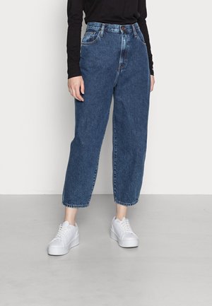Personne portant un jean denim bleu taille haute avec des jambes légèrement effilées et des baskets blanches à plateforme, debout sur un sol gris.